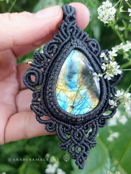 Close-up of a teardrop labradorite pendant with intricate black macrame design and vibrant iridescent glow in natural outdoor setting