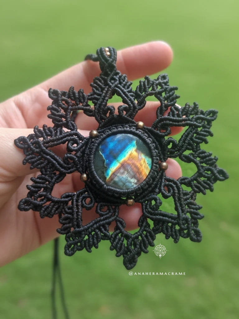 Brown macrame pendant with a colorful stone held in a hand against a green background