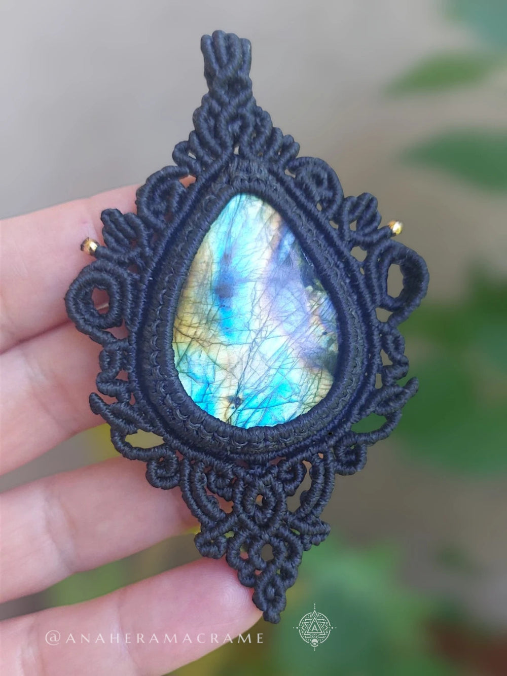 Frontal view of pear-shaped labradorite pendant framed in ornate macrame, held up against blurred natural scenery