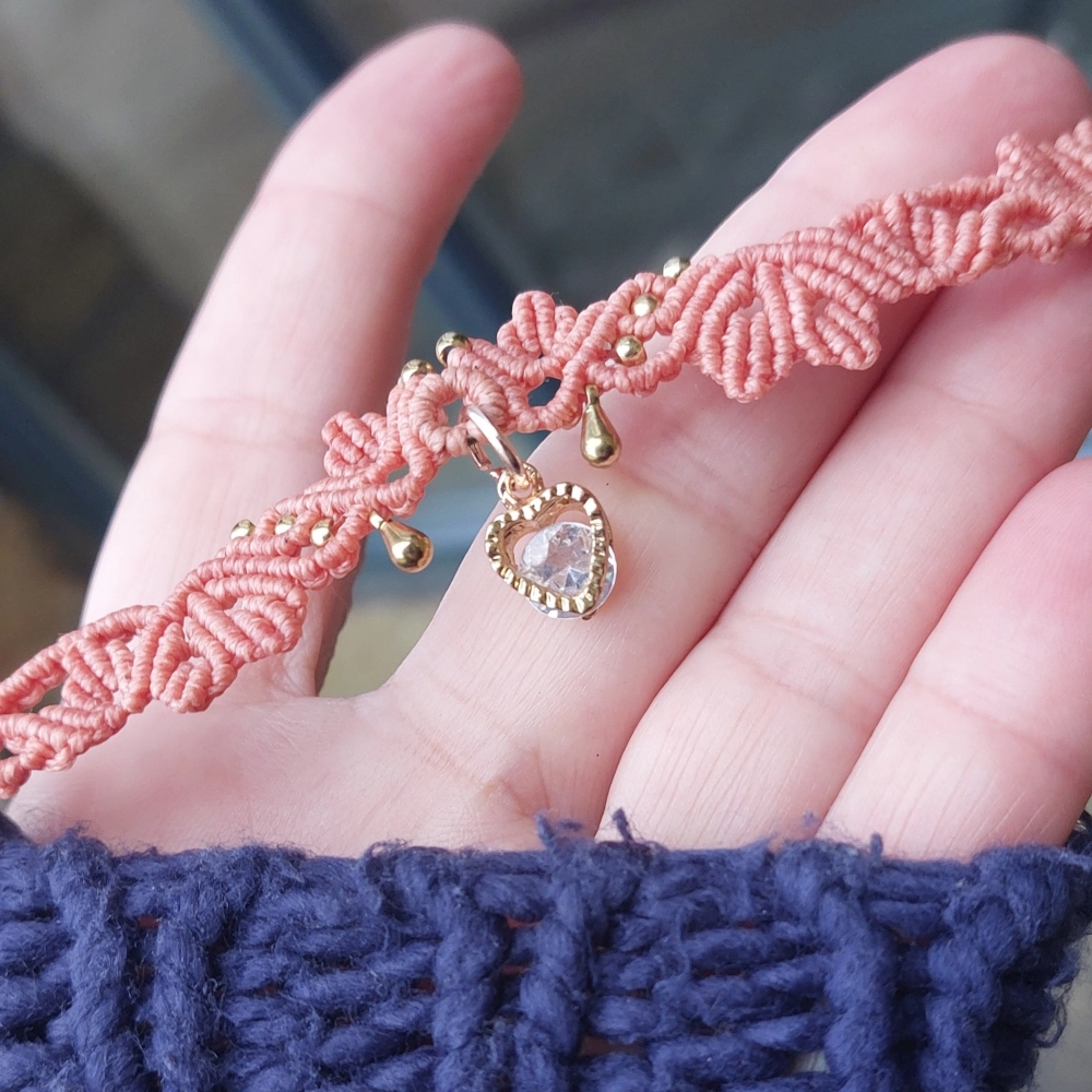 Handcrafted macrame choker featuring a soft coral-pink hue, a Cubic Zirconia heart charm centerpiece, and elegant golden accents.
