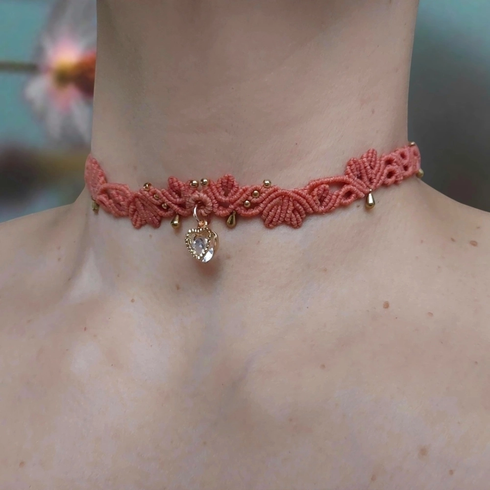 Handmade coral macrame choker featuring a Cubic Zirconia heart charm and golden accents, adjustable with a sliding lock for a custom fit.
