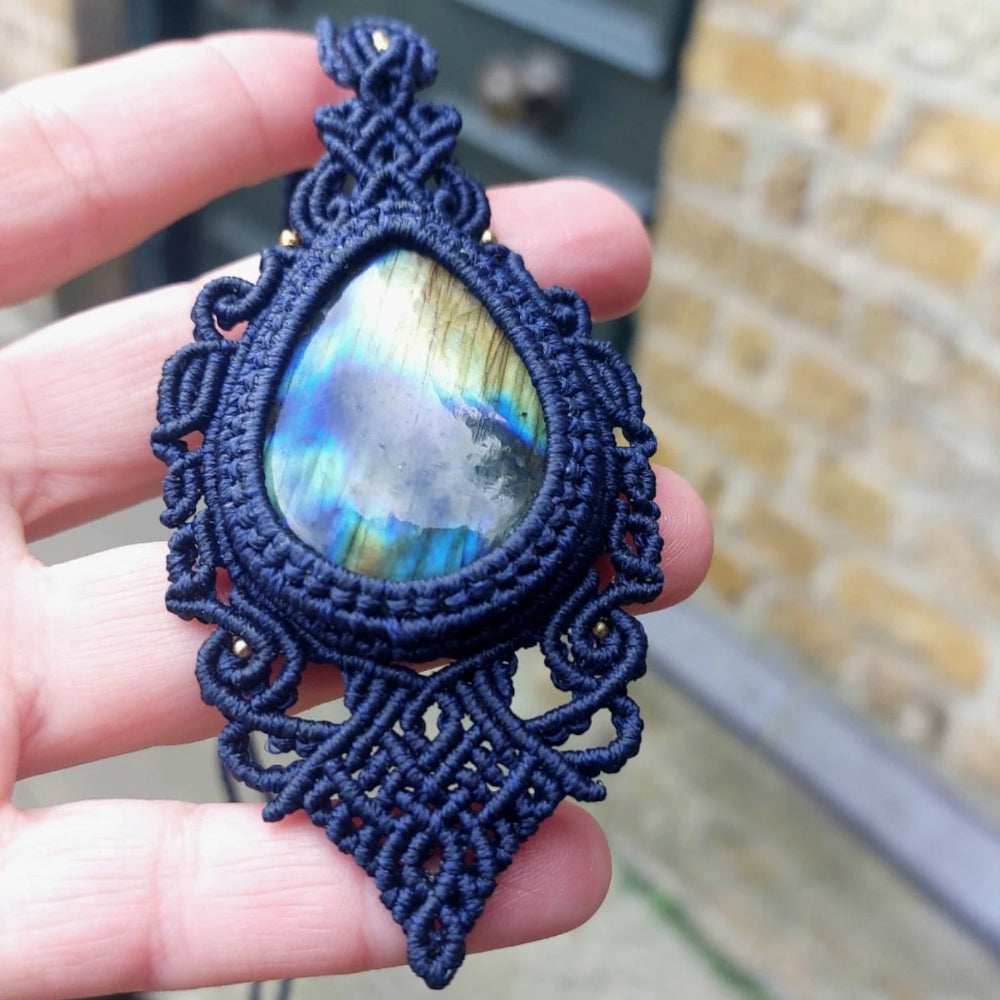 Labradorite stone pendant glowing with internal light, set in an adjustable blue macramé cord necklace, held outdoors.