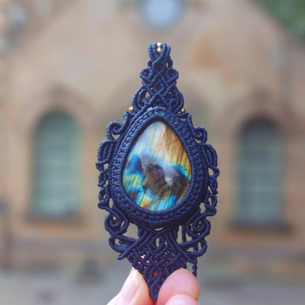 Hand holding a teardrop labradorite pendant with vibrant blue and gold flashes, encased in hand-knotted blue macramé.

