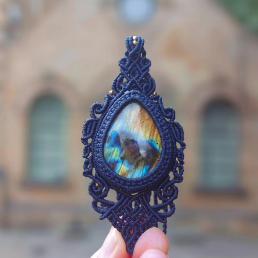 Hand holding a teardrop labradorite pendant with vibrant blue and gold flashes, encased in hand-knotted blue macramé.

